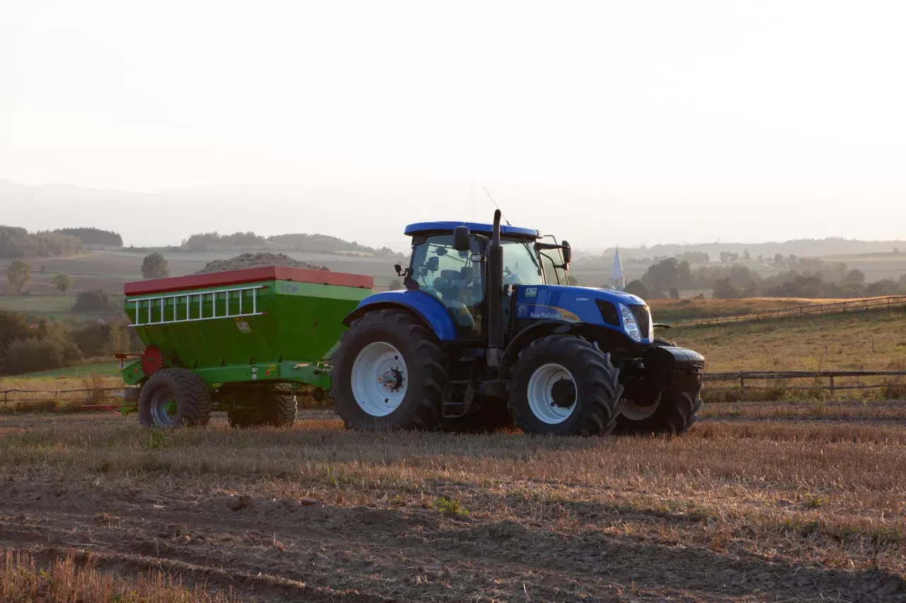 Niebieski traktor z przyczepą nawozową, gotowy do pracy na polu. Rolnik zastanawia się, jakie wapno na dofinansowanie wybrać.