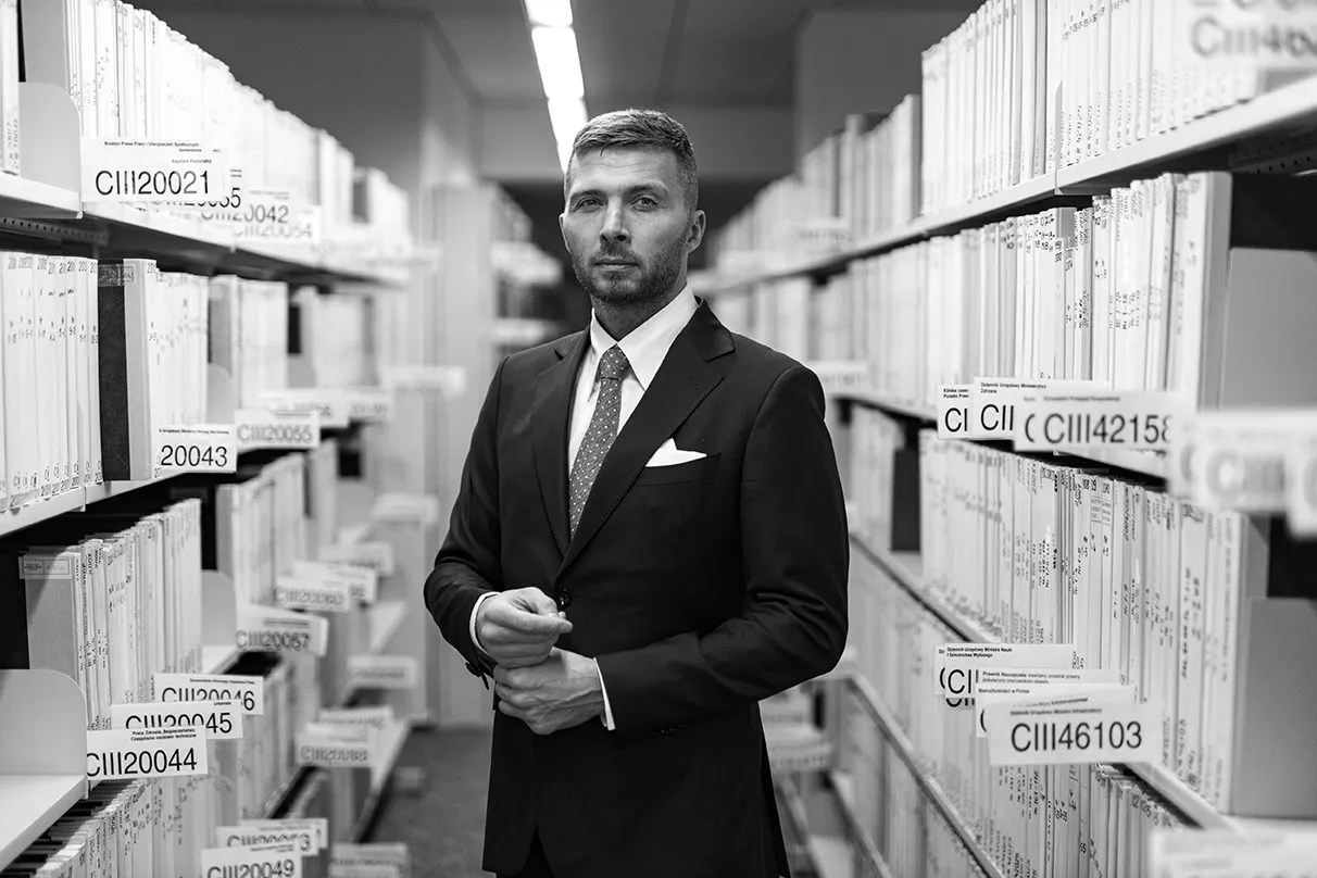 Mężczyzna w garniturze stoi w archiwum, wśr&oacute;d regał&oacute;w z dokumentami. Może to być związane z nowelizacją kodeksu karnego.