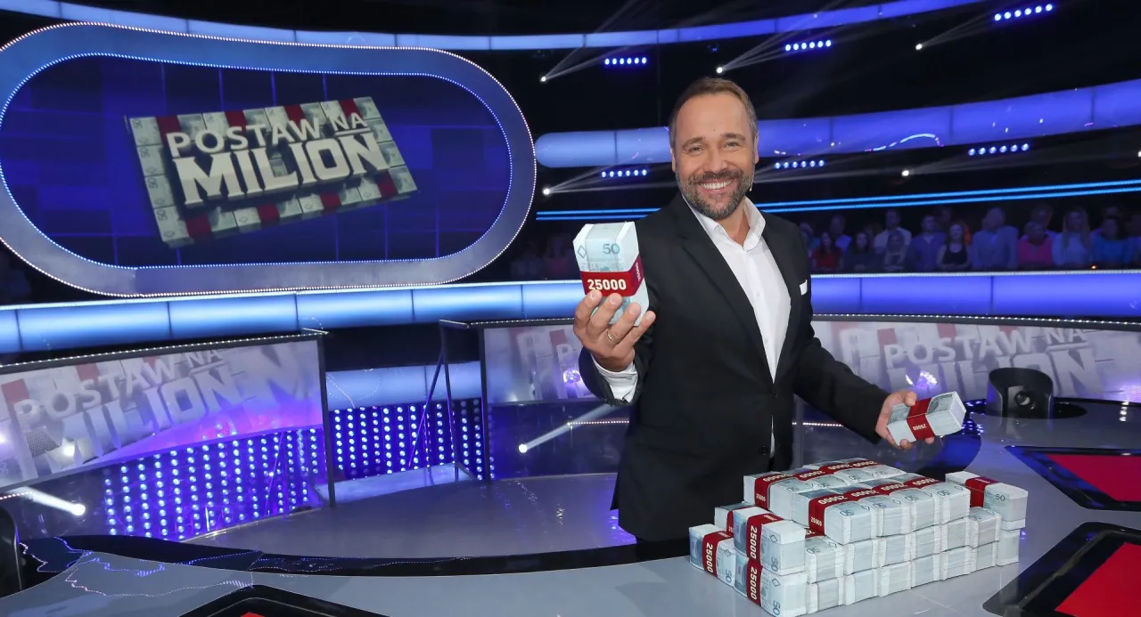 Uśmiechnięty prowadzący w garniturze trzyma stos banknot&oacute;w. Chcesz wiedzieć, jak dostać się do programu Milionerzy?