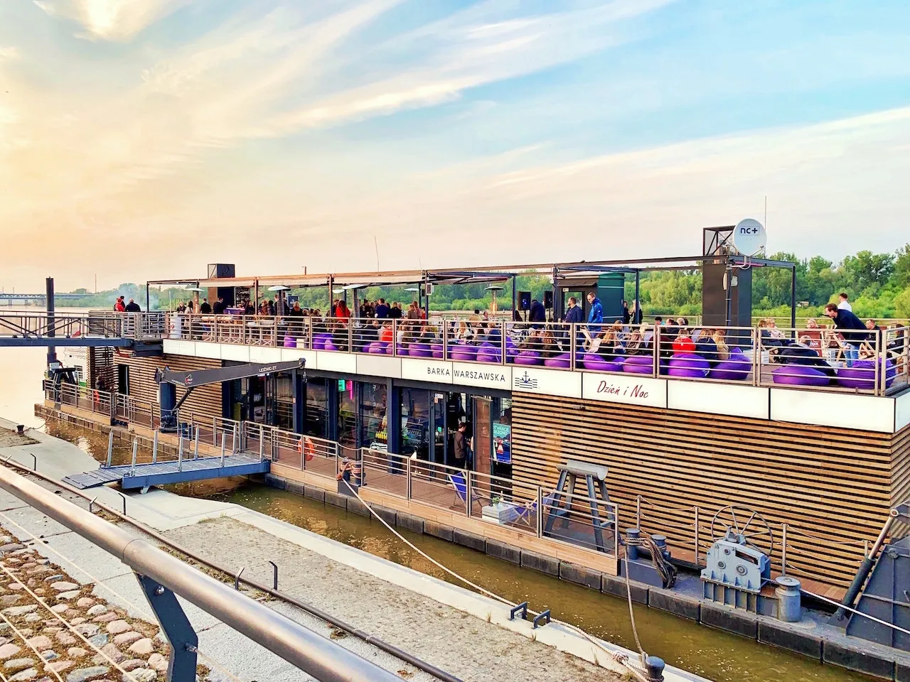 Barka warszawska Dzień i Noc tętni życiem. Ludzie relaksują się na fioletowych pufach na górnym pokładzie, ciesząc się widokiem Wisły.