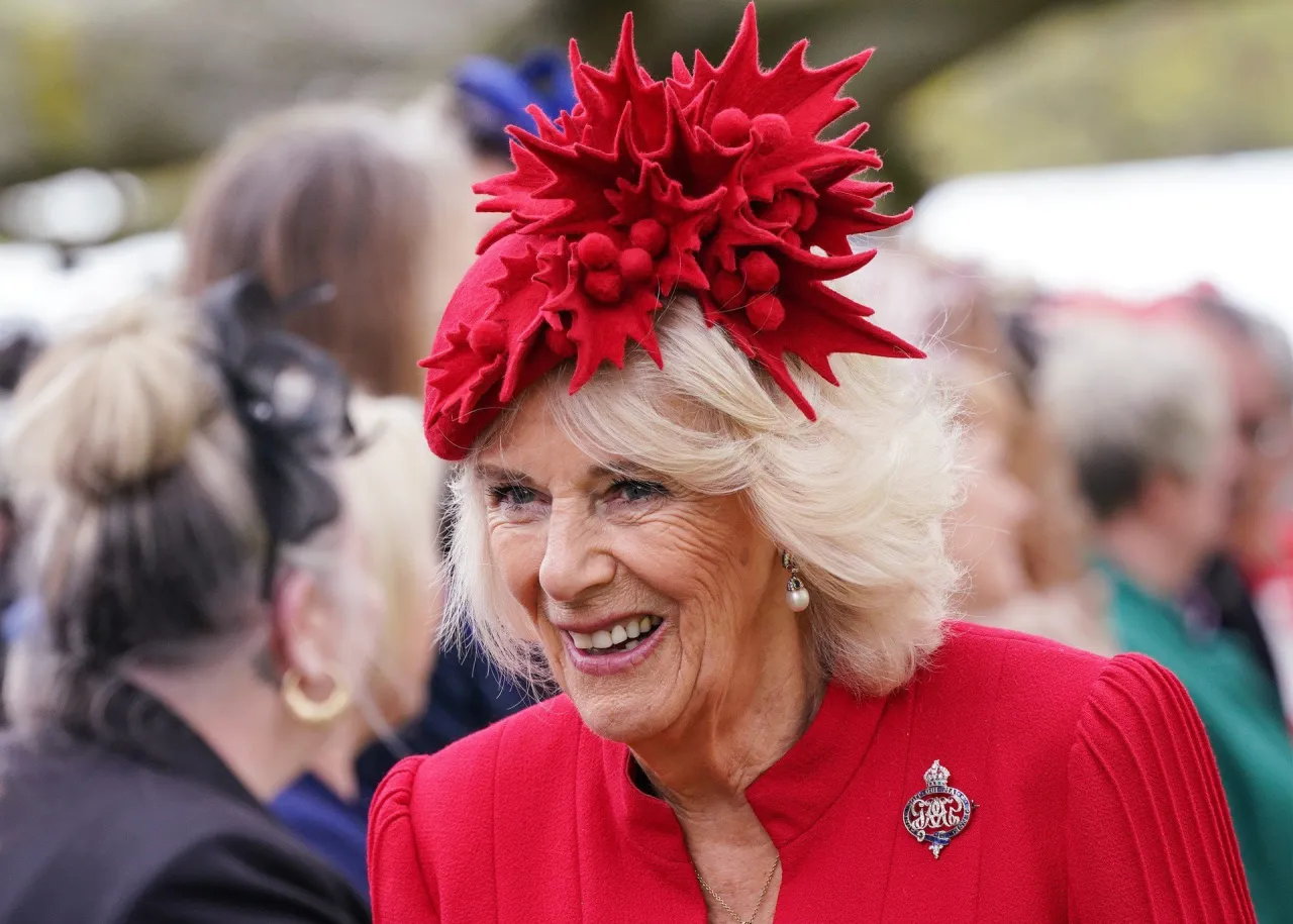 Uśmiechnięta Camilla Parker Bowles w czerwonym kapeluszu z ozdobnym kwiatem i broszką.