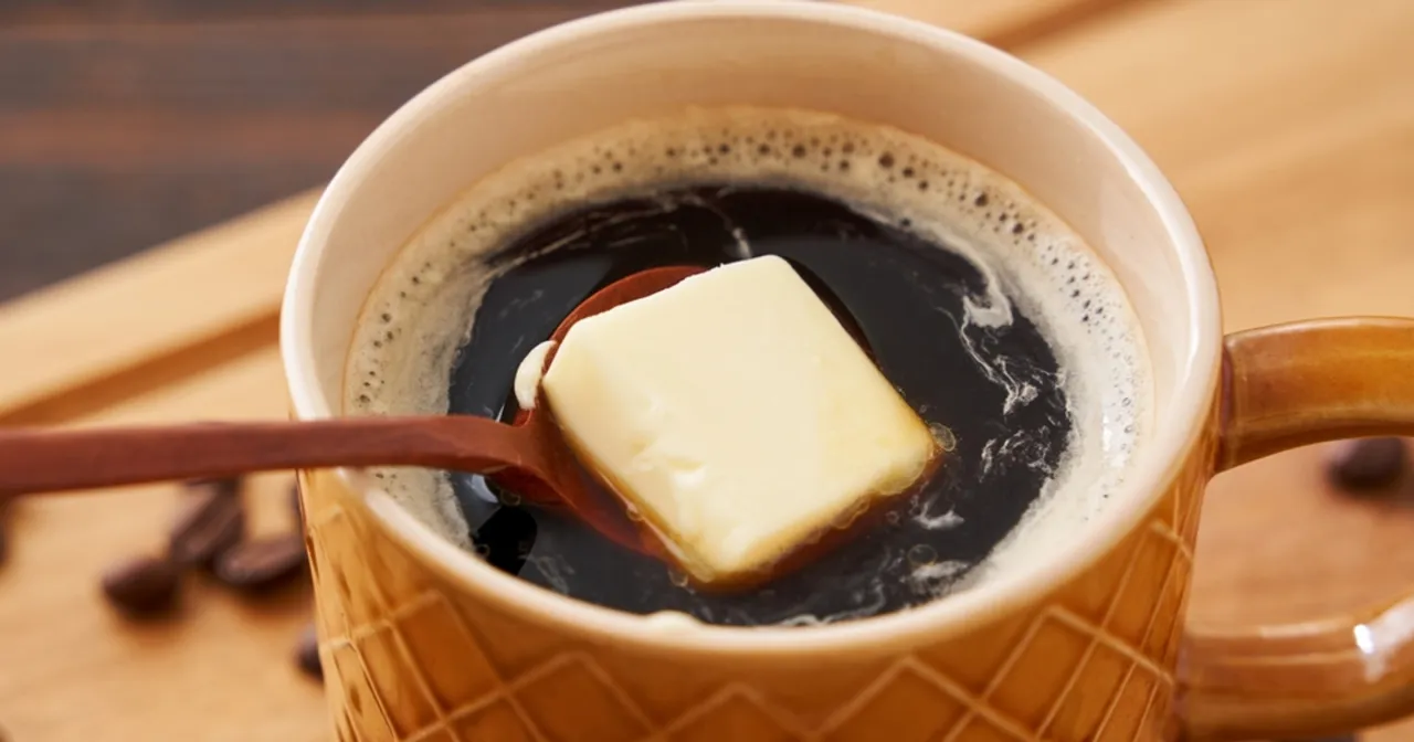 Kawa z masłem na odchudzanie w brązowym kubku, z drewnianą łyżeczką trzymającą kostkę masła.