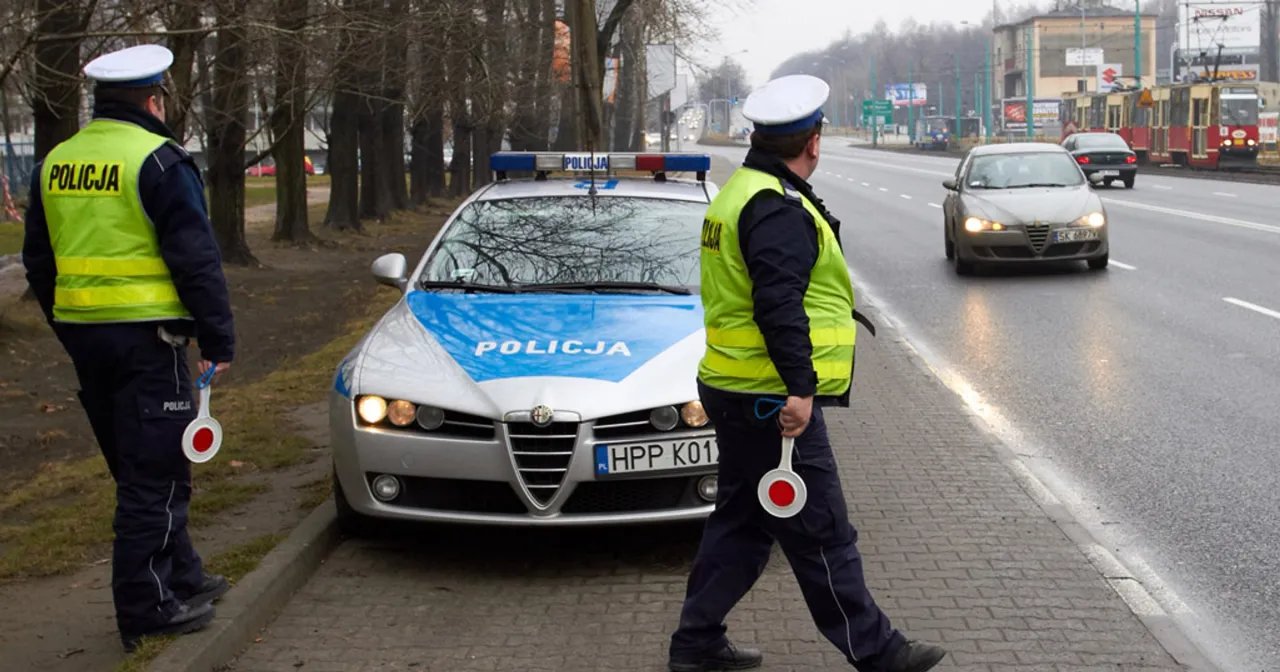 Policjanci zatrzymują kierowcę z powodu przekroczenia prędkości. Na drodze stoi radiow&oacute;z, a jeden z funkcjonariuszy trzyma lizak do zatrzymywania pojazd&oacute;w.