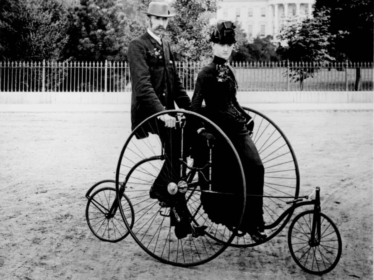 Para, jak powstał rower: mężczyzna i kobieta na welocypedzie, z wielkim przednim kołem i małym tylnym.