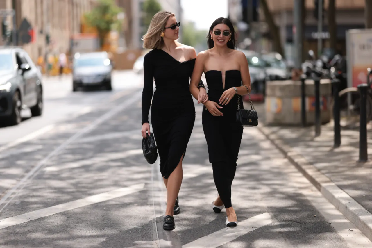 Dwie kobiety w czarnych sukienkach wieczorowych idą ulicą. Pokazują, jak dobrać buty do sukienki wieczorowej, prezentując eleganckie stylizacje.