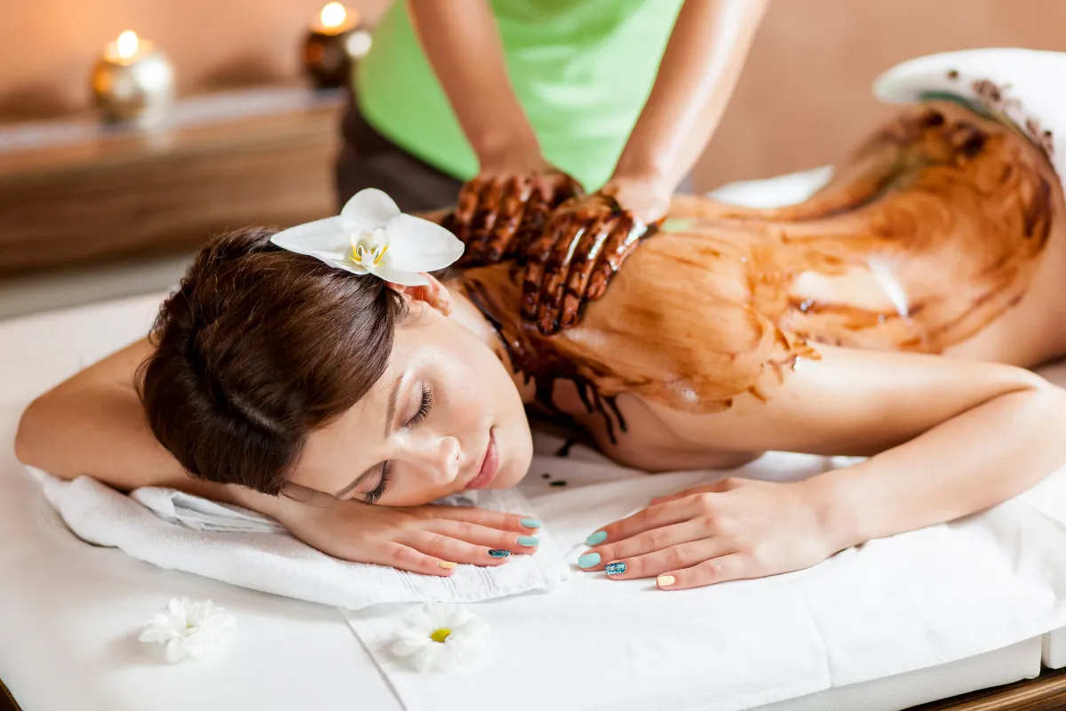 Kobieta leży na stole do masażu, a jej plecy są pokryte czekoladą do masażu spa. Delikatne świece i kwiaty tworzą relaksującą atmosferę.