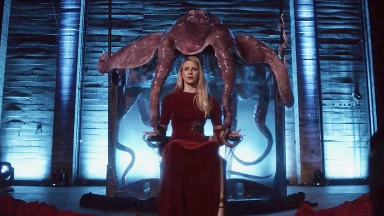 Kobieta w czerwonej sukni siedzi przed ogromnym, przypominającym ośmiornicę stworzeniem. To scena z serialu