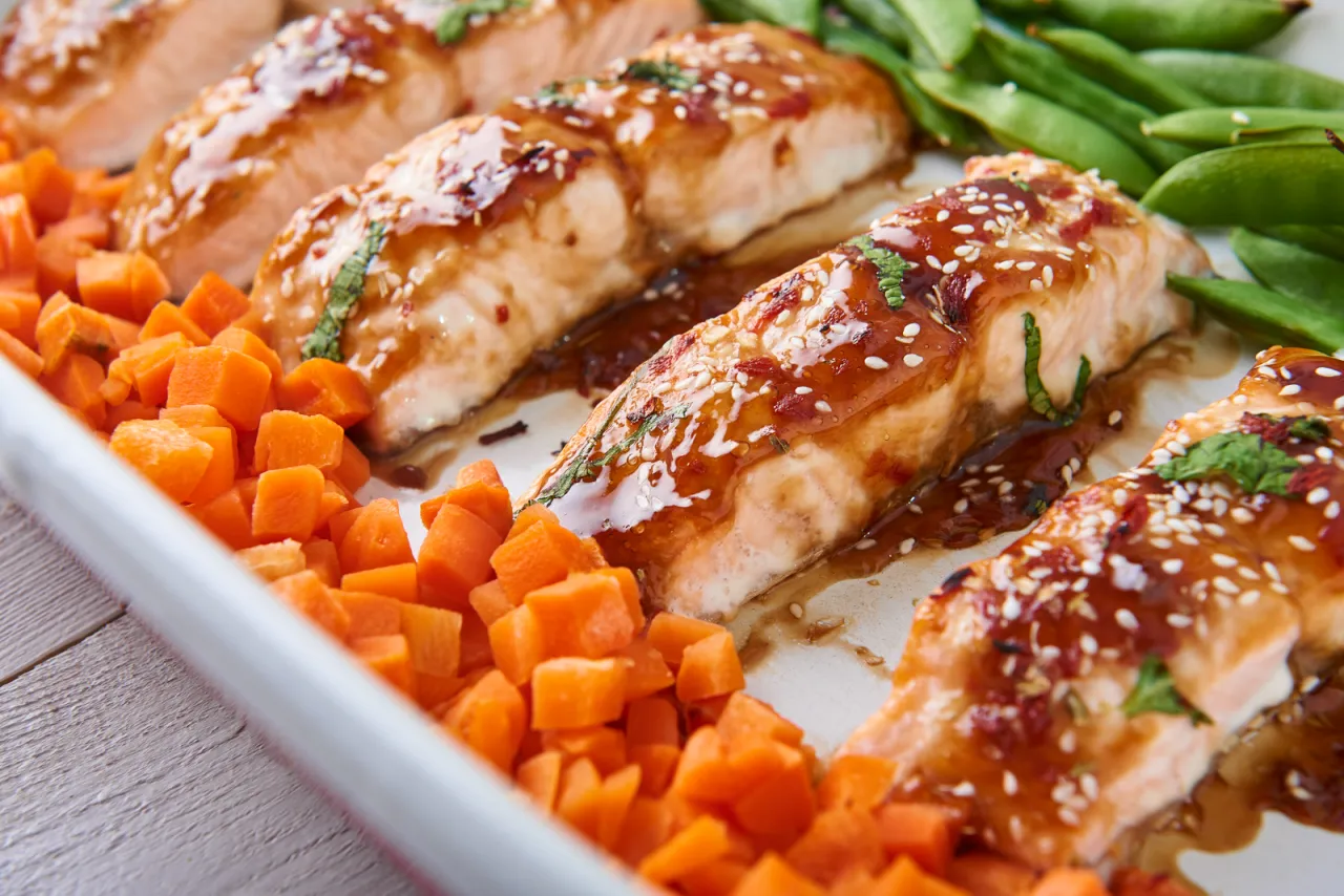 Soczysty łosoś w sosie sojowym, posypany sezamem i ziołami, podany z chrupiącą marchewką i zielonym groszkiem. Pyszne danie gotowe do spożycia.