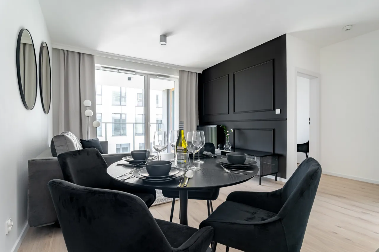 Eleganckie mieszkanie inwestycyjne co to znaczy? Stylowy salon z jadalnią, czarny stół, welurowe krzesła, butelka wina i widok na nowoczesny budynek.