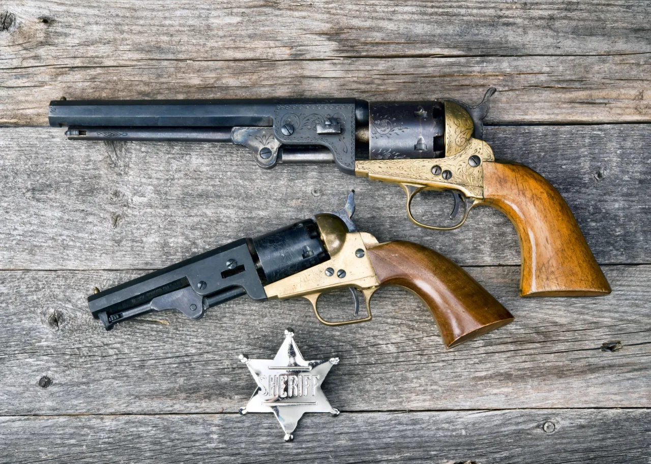 Dwa zabytkowe rewolwery i odznaka szeryfa na drewnianym tle. Czy myśliwy może kupić czarny proch do takich broni?