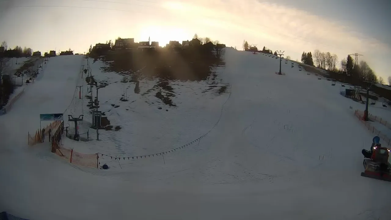 Ośnieżony stok narciarski na olczańskim wierchu z wyciągami i domkami w tle. Na dole pracuje ratrak.
