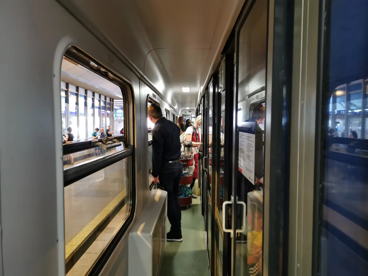 Wnętrze pociągu IC. Widać korytarz z drzwiami i oknami, ludzi i w&oacute;zek z przekąskami.