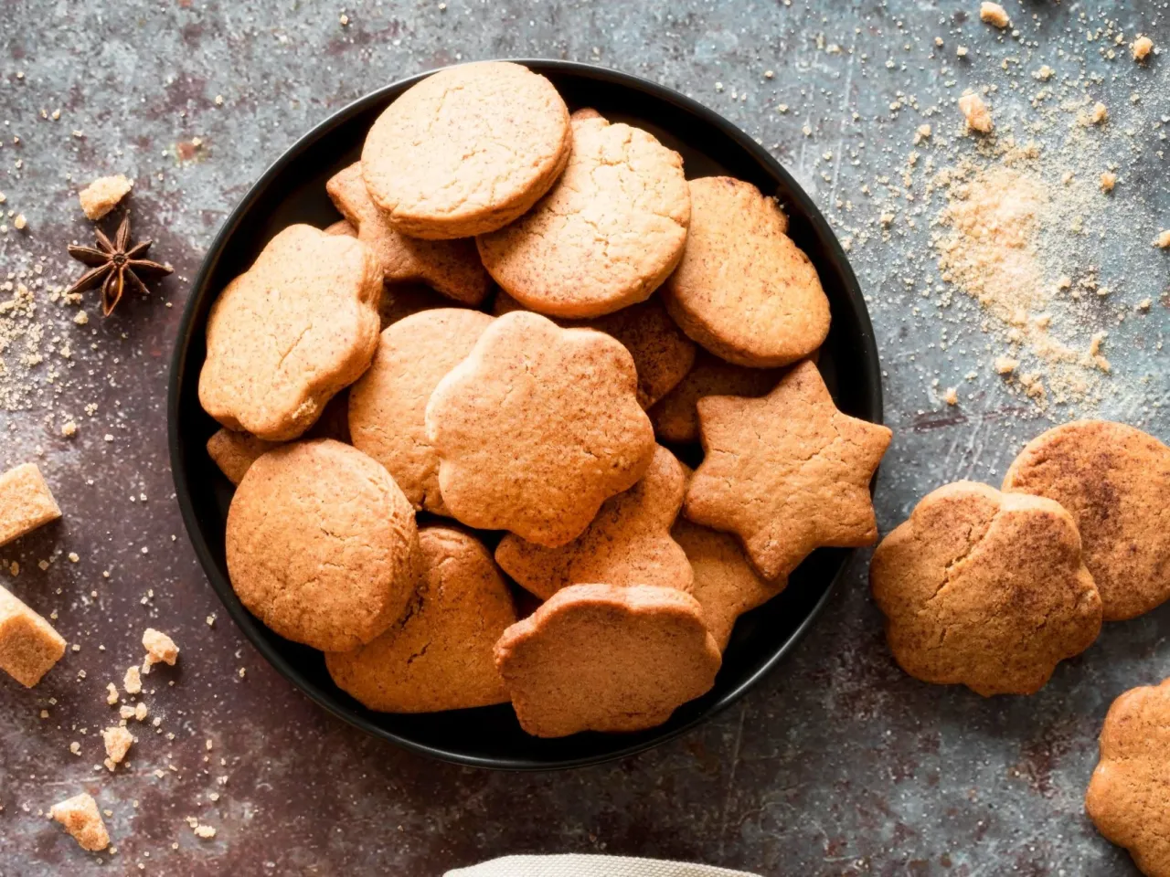 Talerz pełen okrągłych i gwiazdkowych ciasteczek, idealnych do przepisu na zdrowe ciasteczka. Obok rozsypany cukier i anyż.
