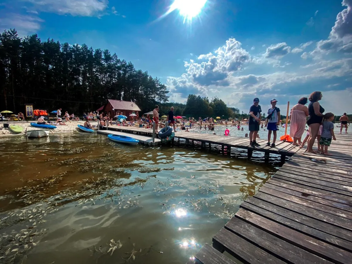 Słoneczny dzień nad zalewem w Janowicach. Ludzie odpoczywają na plaży i spacerują po drewnianym pomoście.
