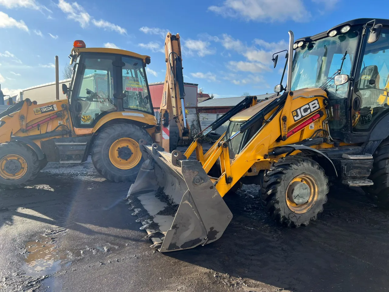 Dwie żółte koparki JCB stoją na błotnistym placu. Jedna z nich ma na szybie napis