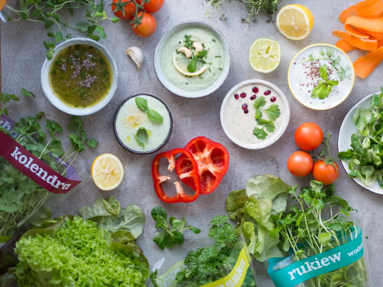 Jak zrobić sos do sałatki z oleju? Na stole świeże zioła, warzywa, cytryny i miseczki z sosami.