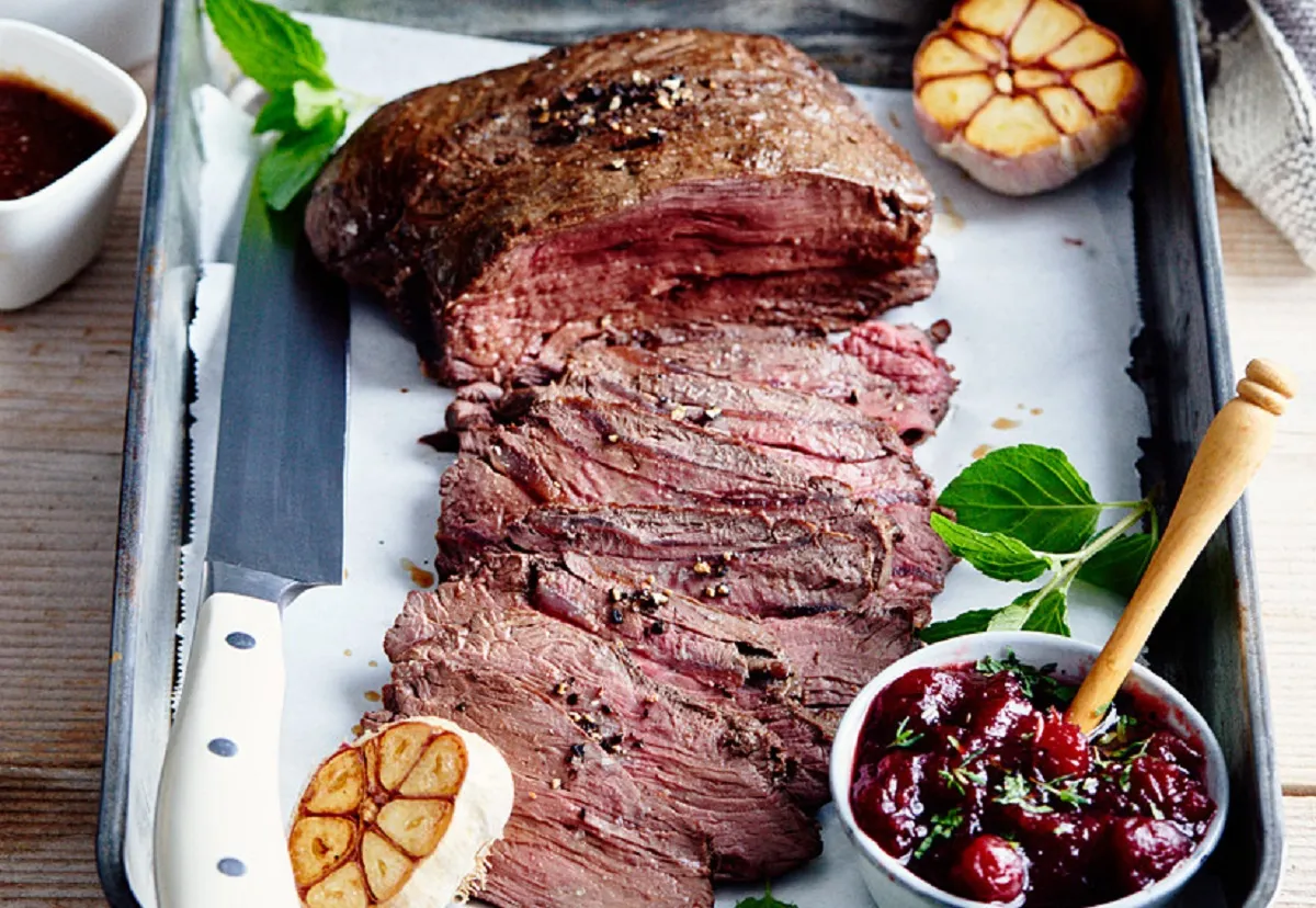 Schab z jelenia, soczysty i idealnie wysmażony, pokrojony w plastry. Obok sos żurawinowy, pieczony czosnek i zioła. Idealny przepis, jak przygotować schab z jelenia.