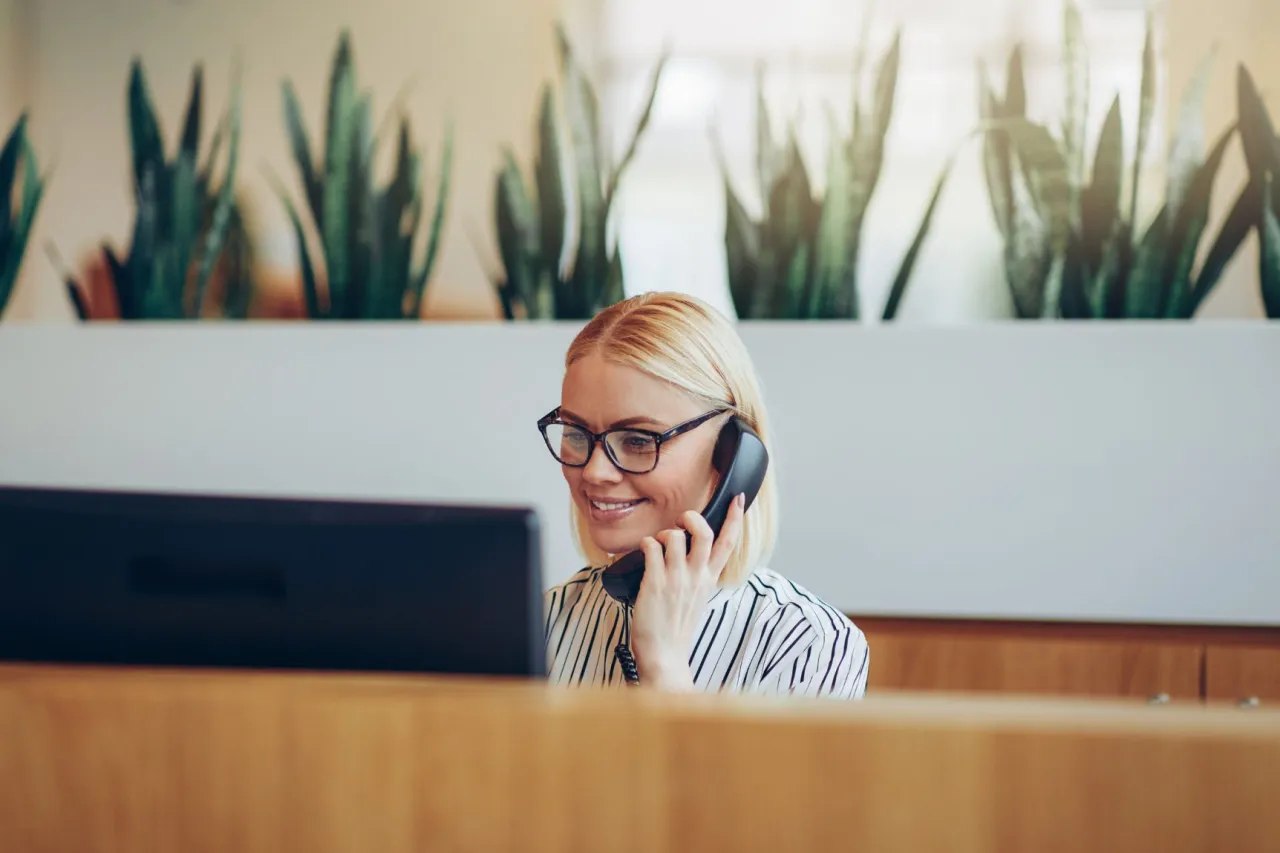 Uśmiechnięta blondynka w okularach rozmawia przez telefon, siedząc przy biurku. Ciekawe, ile zarabia recepcjonista na takim stanowisku.