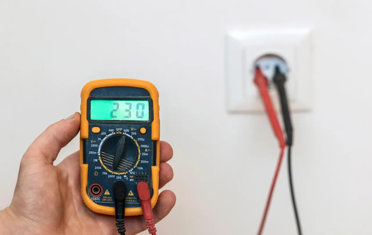 Multimetr pokazuje 230V, jednostka napięcia elektrycznego mv. W tle gniazdko z sondami.