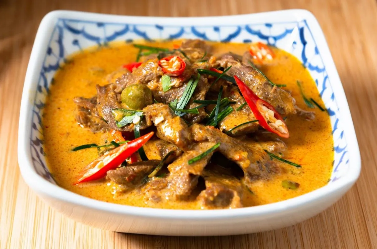 Pyszne tajskie jedzenie: wołowina w kremowym, pomarańczowym sosie curry, ozdobiona plasterkami chili i zielonymi ziołami.