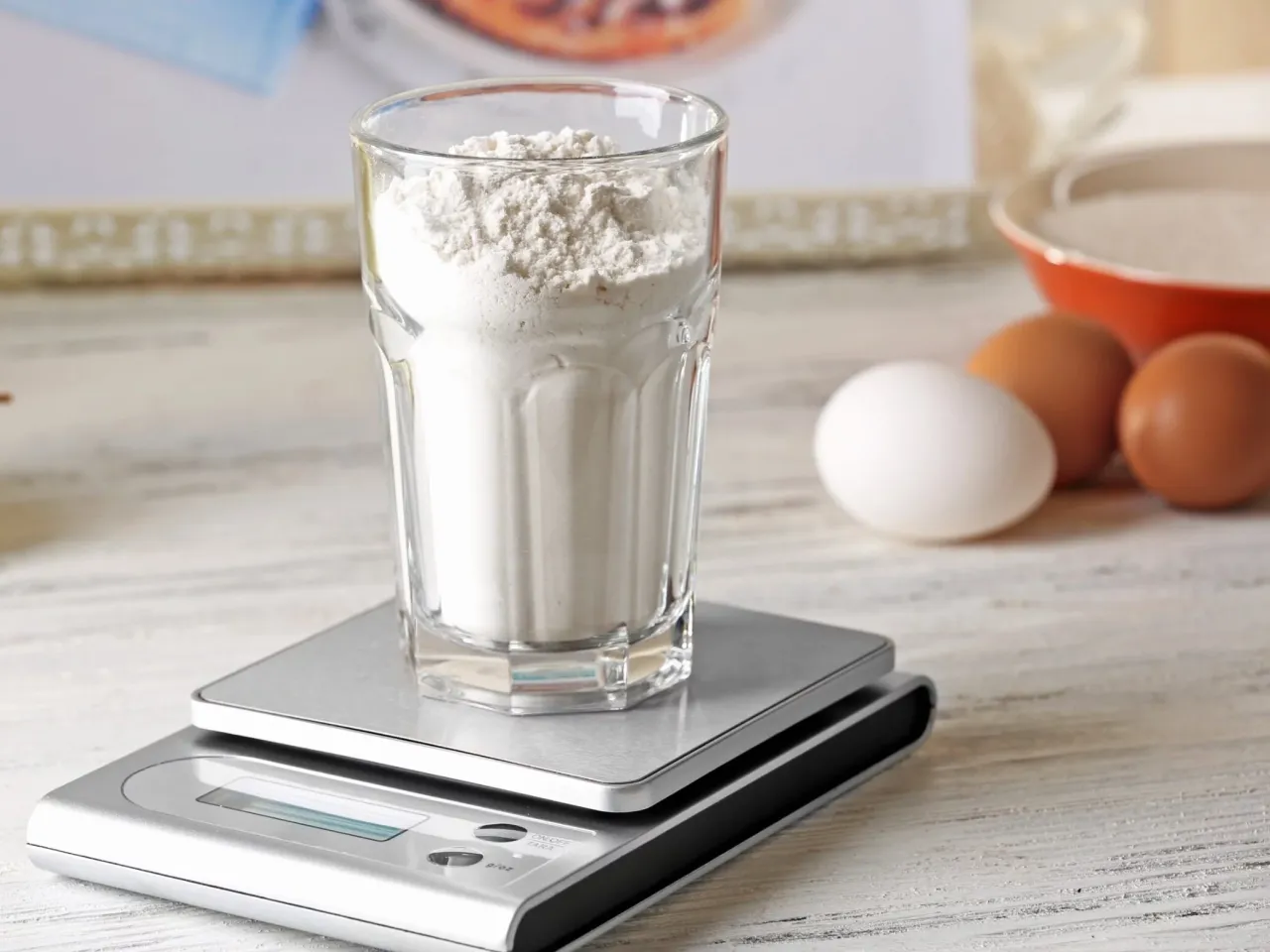 Szklanka mąki na wadze, obok jajka i miseczka z cukrem. Dokładne odmierzanie składników, by wiedzieć, ile g ma łyżka mąki.