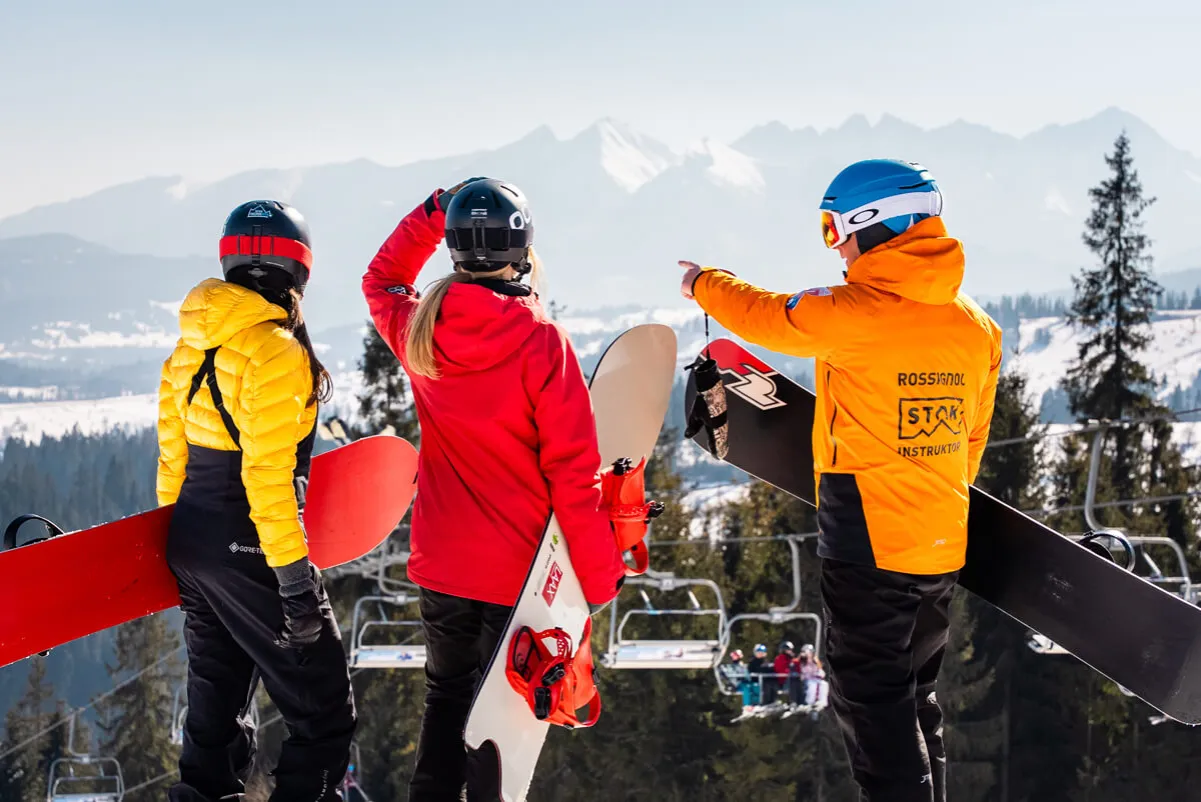 Trzy osoby w zimowych ubraniach stoją na stoku z deskami snowboardowymi. Instruktor w pomarańczowej kurtce wskazuje kierunek. To jest snowboard.