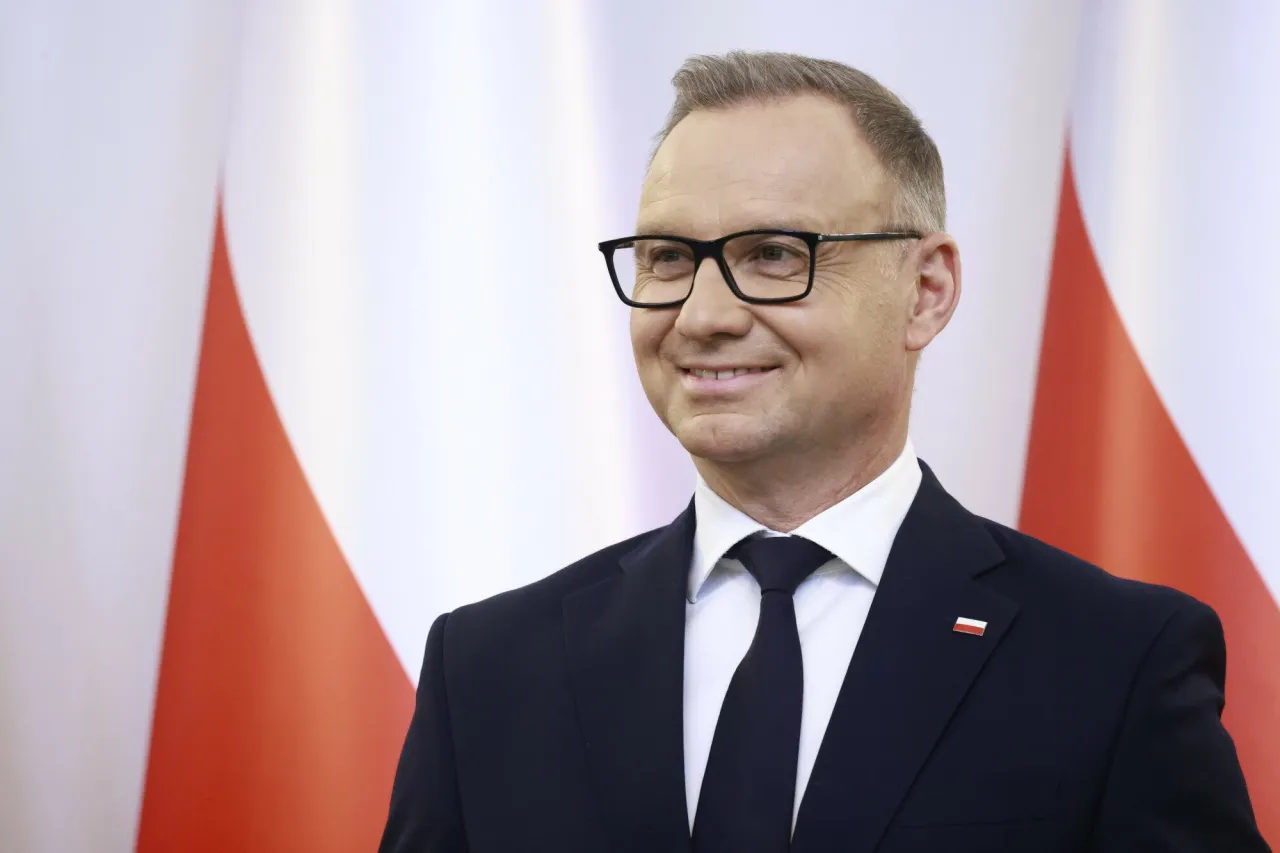 Uśmiechnięty mężczyzna w okularach, w garniturze z przypinką polskiej flagi. W tle biało-czerwona flaga. Ile lat ma Duda?