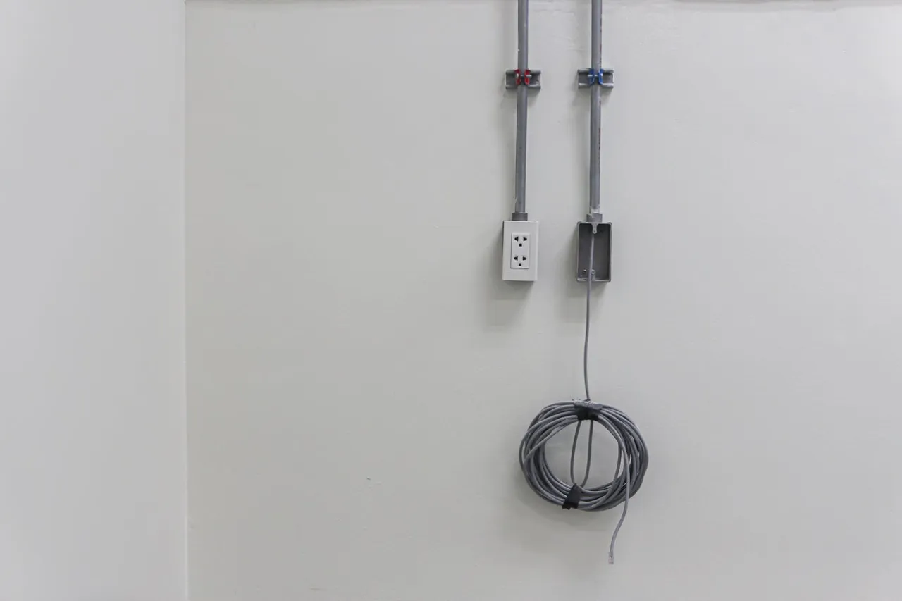 Przewody elektryczne prowadzone w peszlach, przymocowane do ściany. Obok gniazdko i zwinięty kabel, pokazujące jak przymocować kabel do ściany.