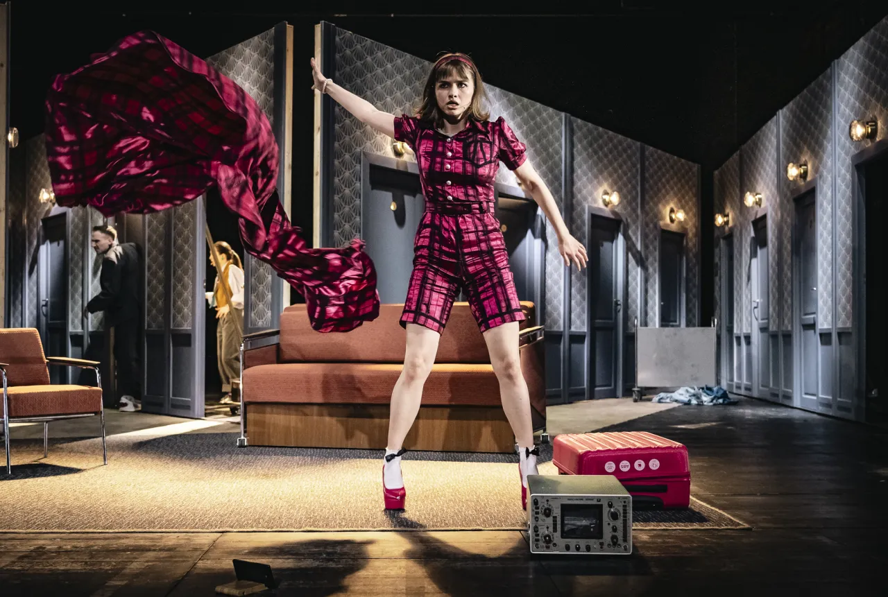 Młoda kobieta w kraciastym stroju, z rozwianą czerwoną tkaniną, pokazuje, o co biega spektakl.