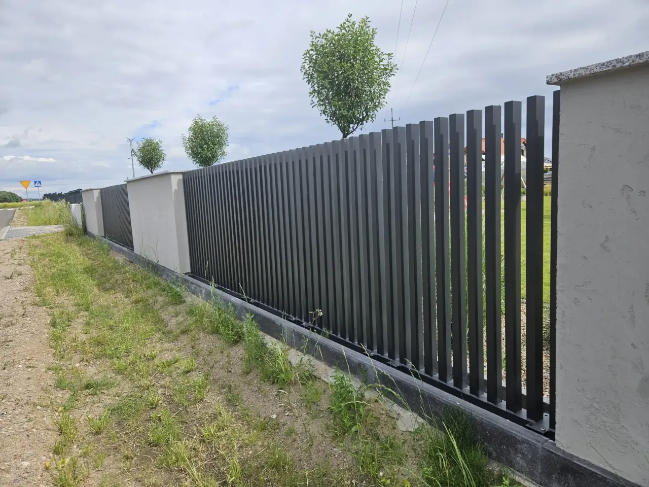 Nowoczesny montaż ogrodzenia panelowego z pionowych, ciemnych profili, osadzonych w betonowych słupkach.
