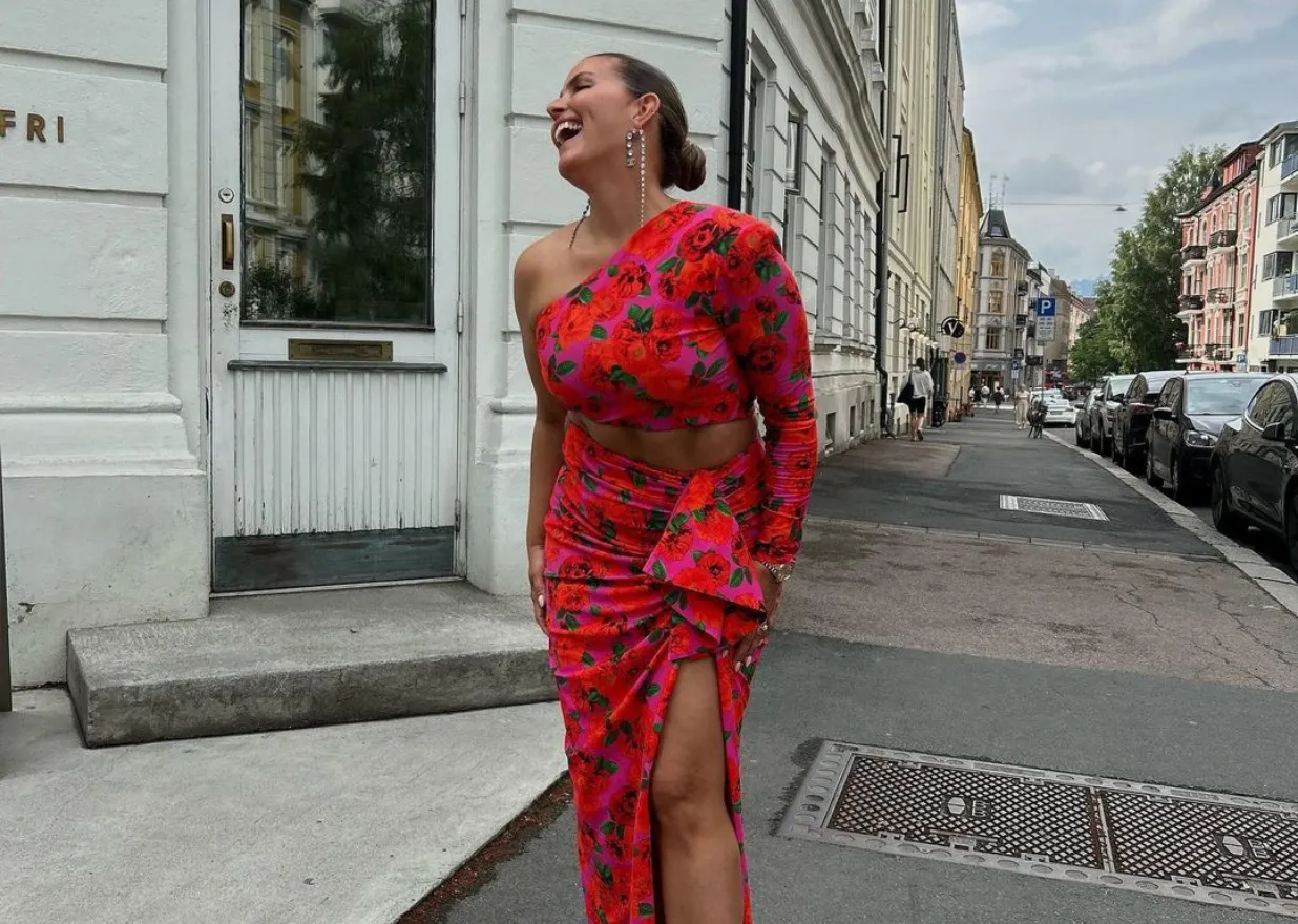 Uśmiechnięta kobieta w kwiecistej sukience na jedno ramię, z rozcięciem i falbaną, prezentuje swoją fryzurę.