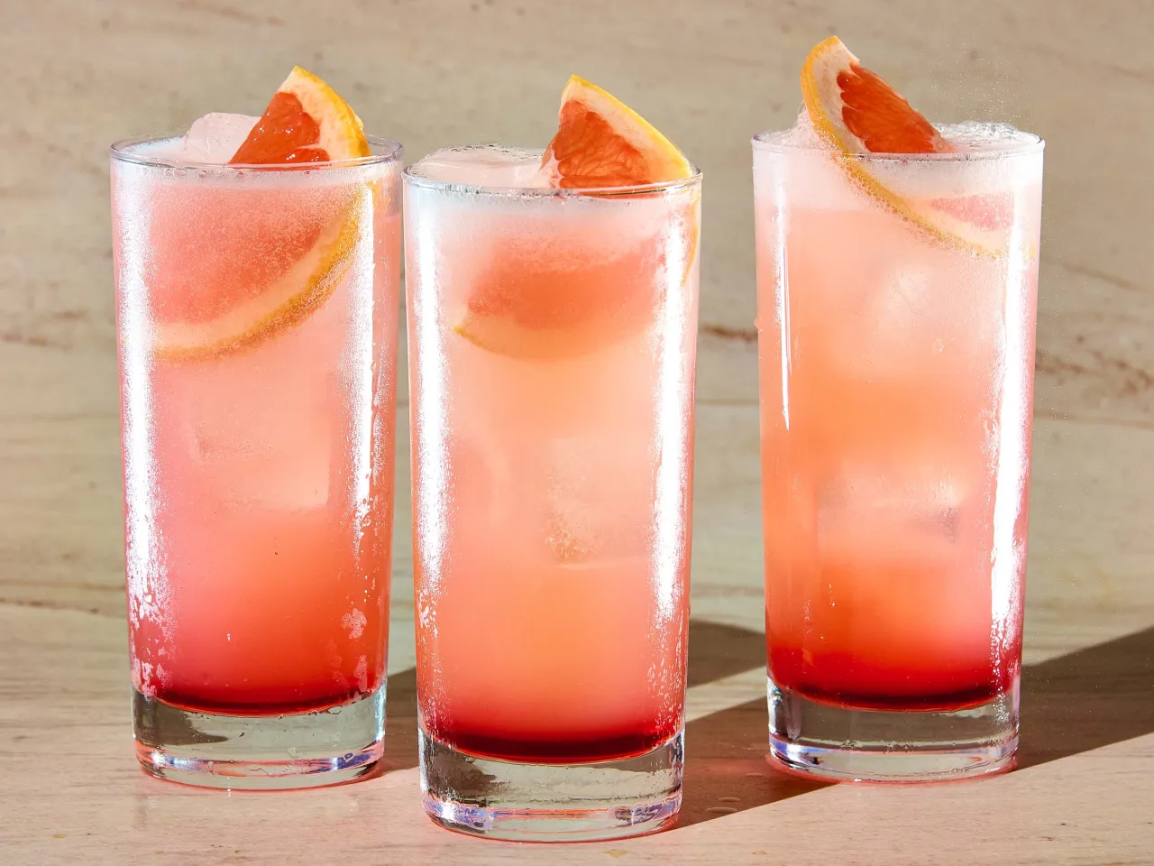 Trzy orzeźwiające drinki: Tequila Sunrise i Paloma, z lodem i plasterkami grejpfruta, na drewnianym blacie.