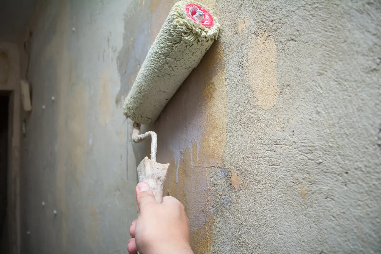 Dłoń trzymająca wałek malarski, który właśnie zaczyna malować chropowatą, szarą ścianę. Zastanawiasz się, ile razy malować ściany, by uzyskać idealny efekt?