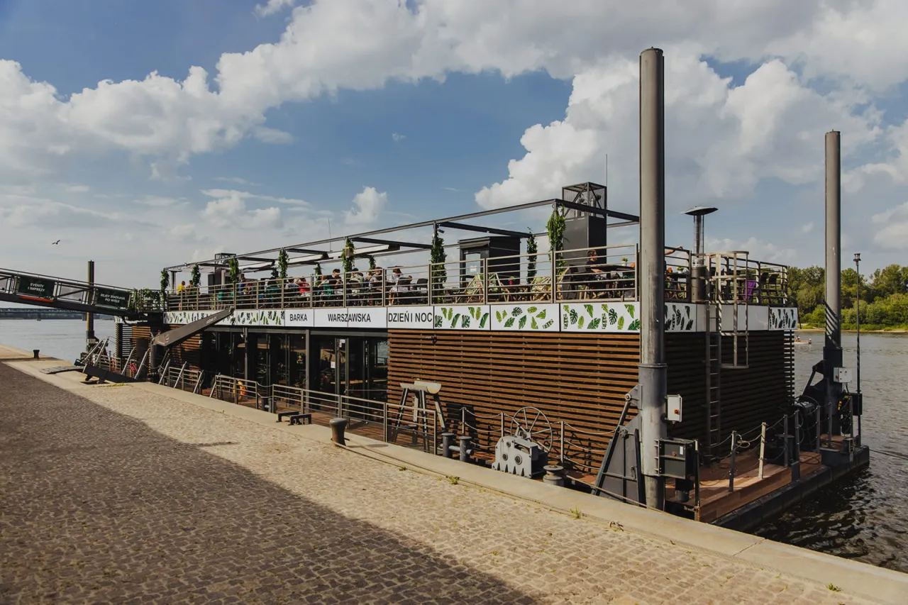 Barka Warszawska Dzień i Noc, pływająca restauracja z ludźmi na pokładzie, cumuje przy nabrzeżu.