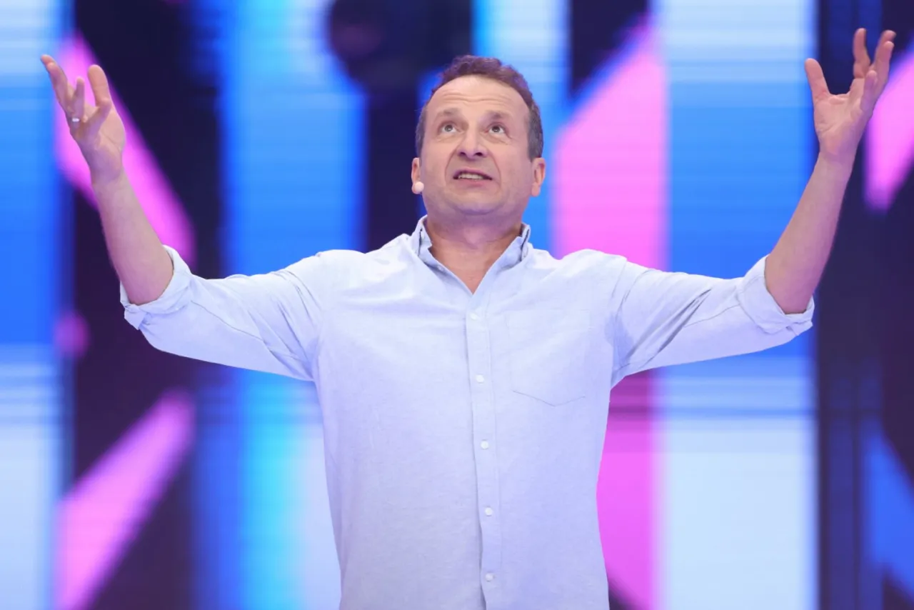 Aktor z Kabaretu Moralnego Niepokoju w błękitnej koszuli, z rękami rozłożonymi na boki, w trakcie jednego ze skeczy.