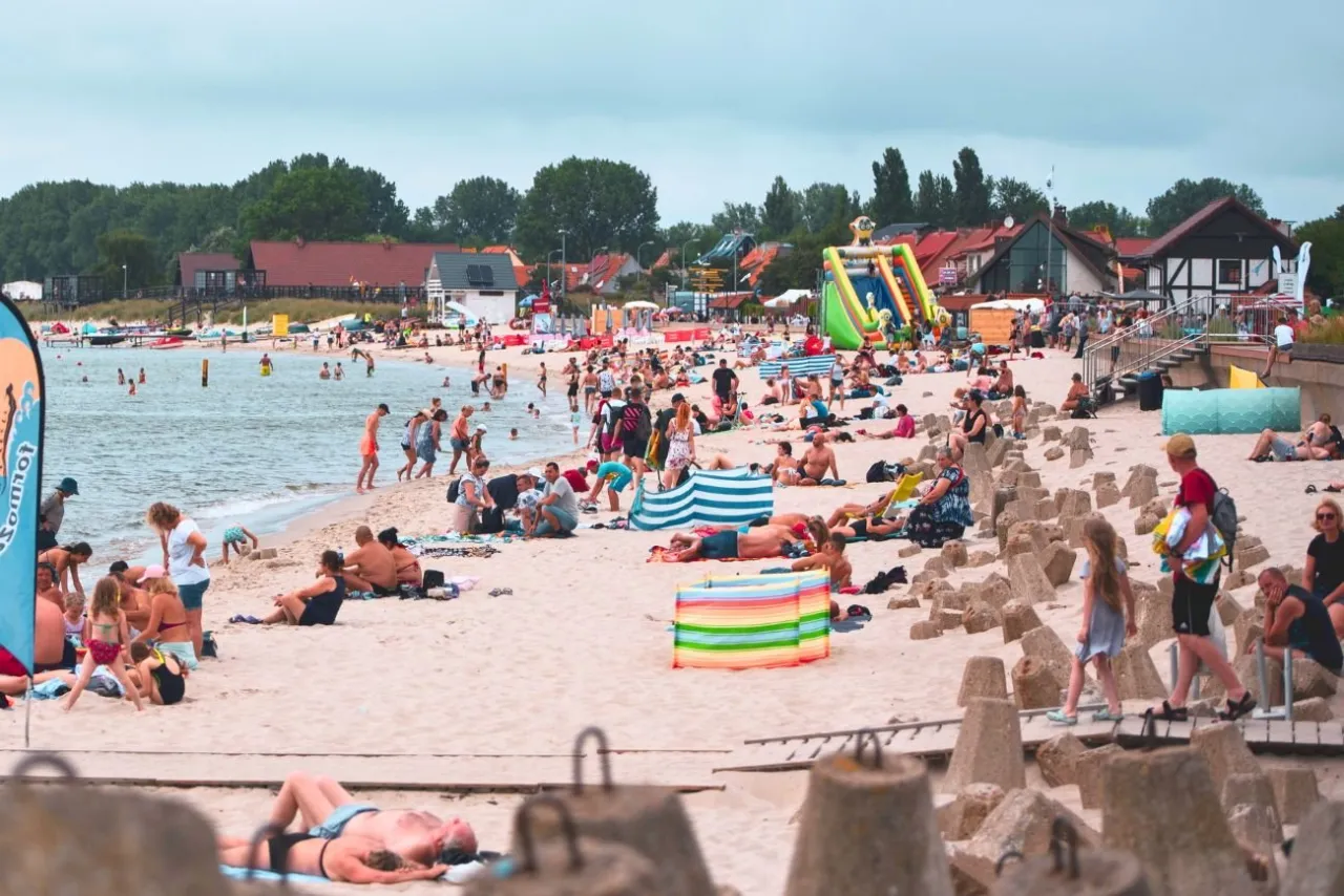 Tłum ludzi odpoczywa na piaszczystej plaży w jednym z nadmorskich miast. W tle widać budynki i zjeżdżalnię dmuchaną.