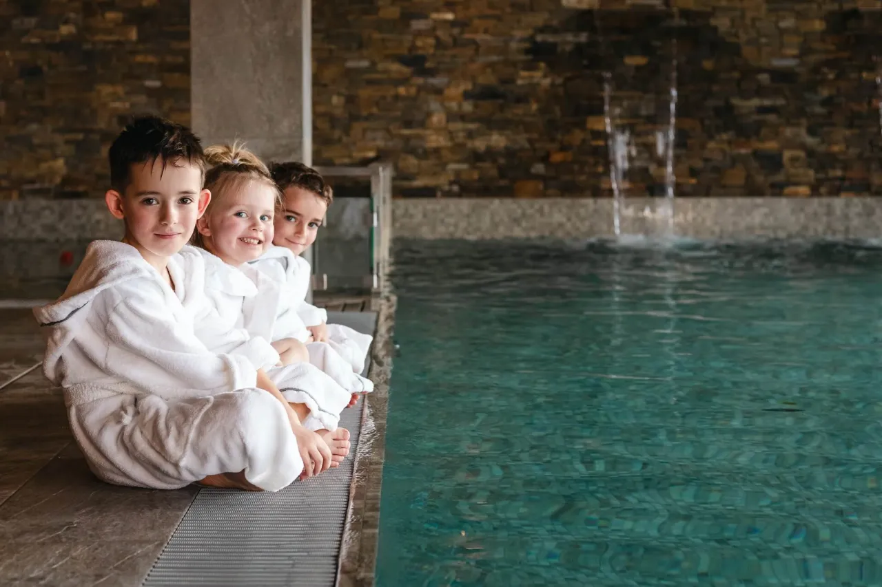 Trójka dzieci w białych szlafrokach siedzi przy basenie, gotowa na sylwester z dziećmi.