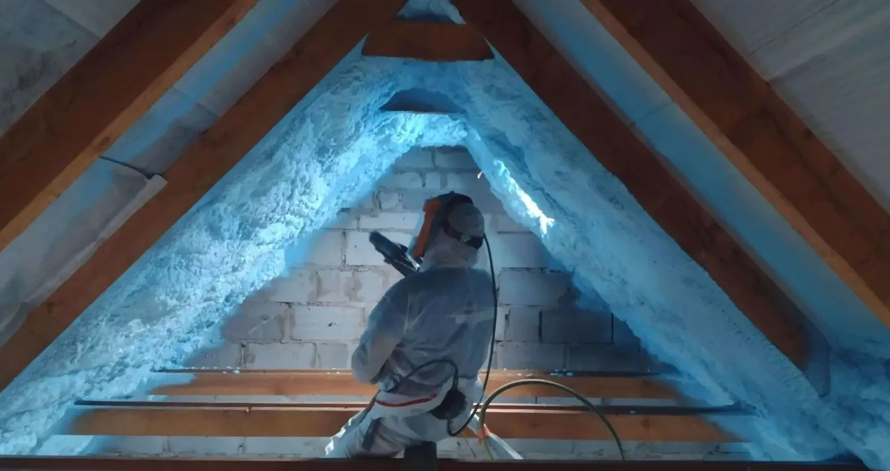 Osoba w kombinezonie natryskuje piankę pur do samodzielnego natrysku na poddasze, tworząc izolację.