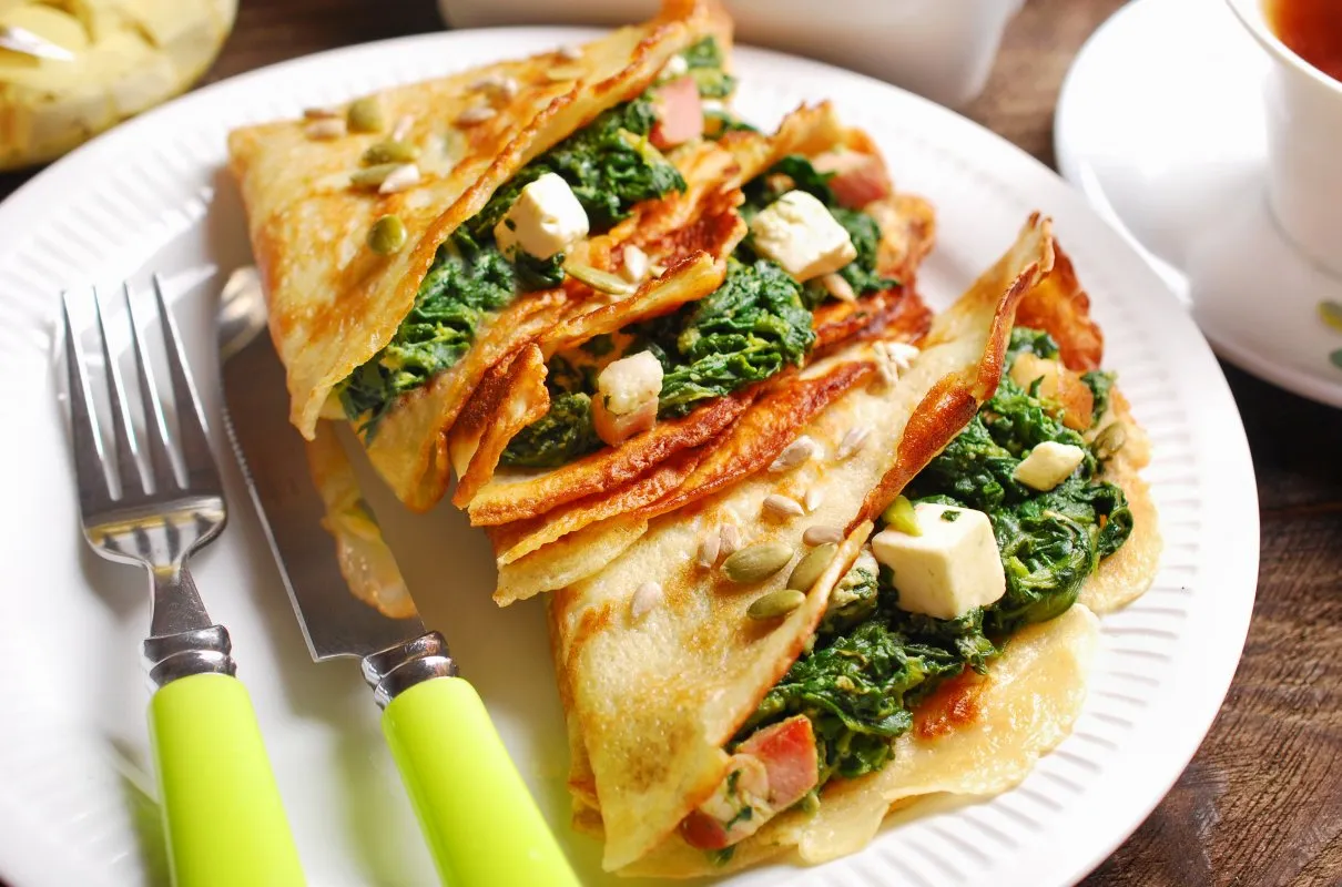 Naleśniki ze szpinakiem i serem feta na obiad, posypane pestkami. Obok widelec i nóż z zielonymi uchwytami.