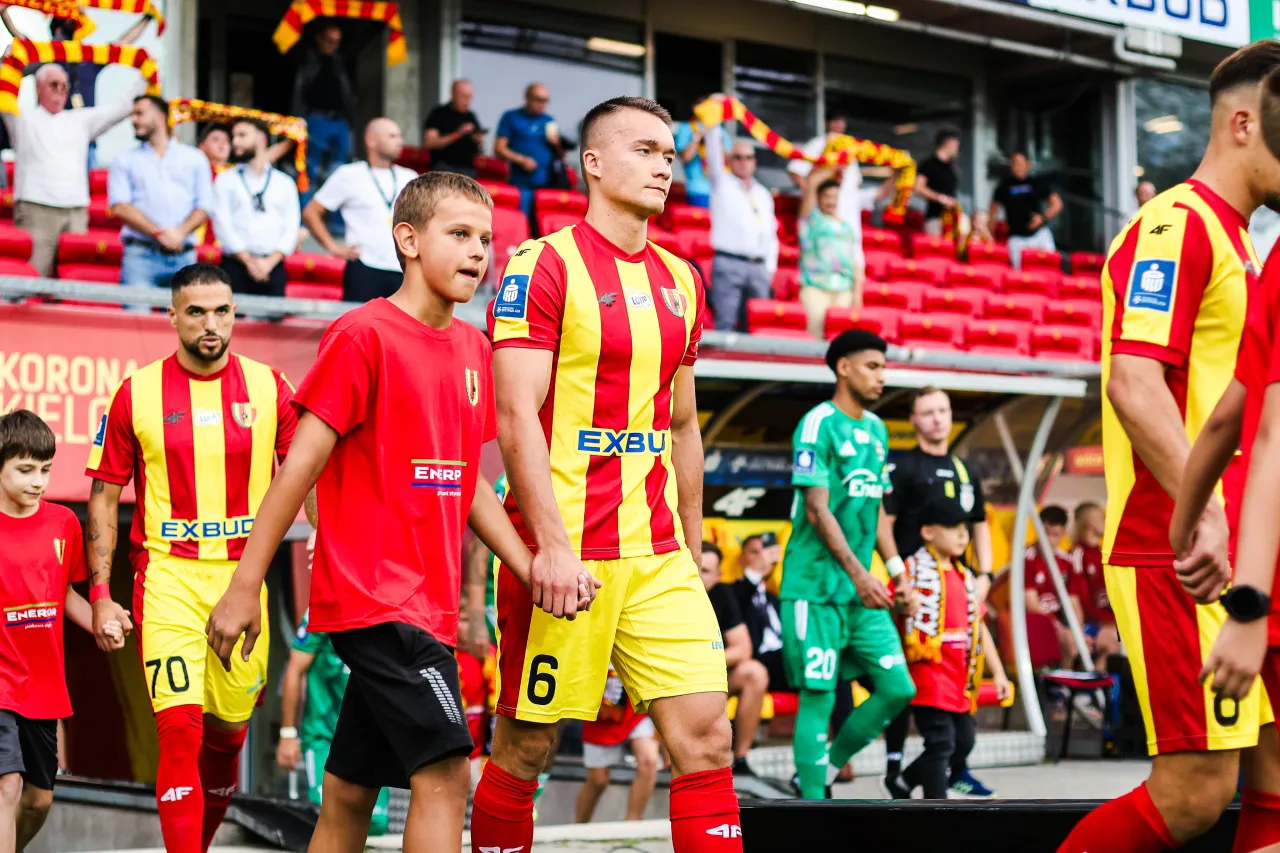 Chłopiec w czerwonej koszulce prowadzi piłkarza w ż&oacute;łto-czerwonym stroju. To może być początek ich drogi, jak zgłosić dziecko do wyprowadzania piłkarzy.