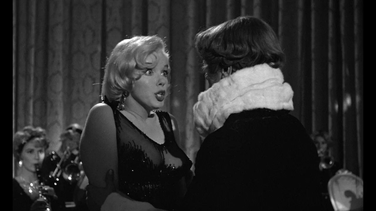 Marilyn Monroe z zaskoczoną miną, w błyszczącej sukni, rozmawia z postacią w futrzanym kołnierzu. Pół żartem pół serio obsada w tle.