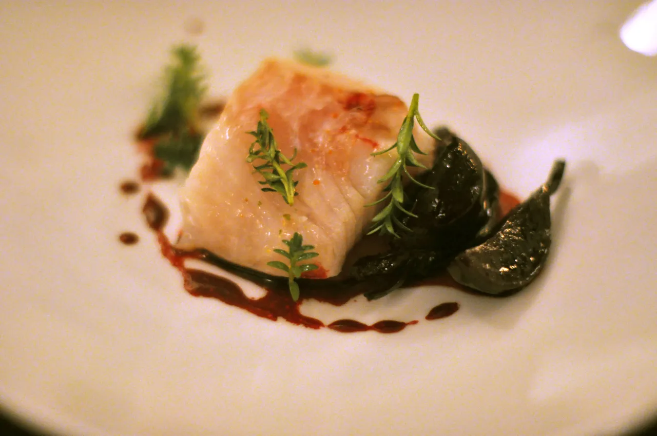 Delikatny kawałek ryby z ciemnymi, błyszczącymi dodatkami i zielonymi gałązkami. Czy ryba jest wegetariańska? To pytanie pozostaje bez odpowiedzi.