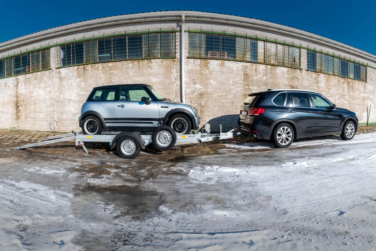 Srebrny Mini Cooper na lawecie, przygotowany do transportu. Obok stoi czarny SUV. Widoczne mocowanie auta na lawecie.