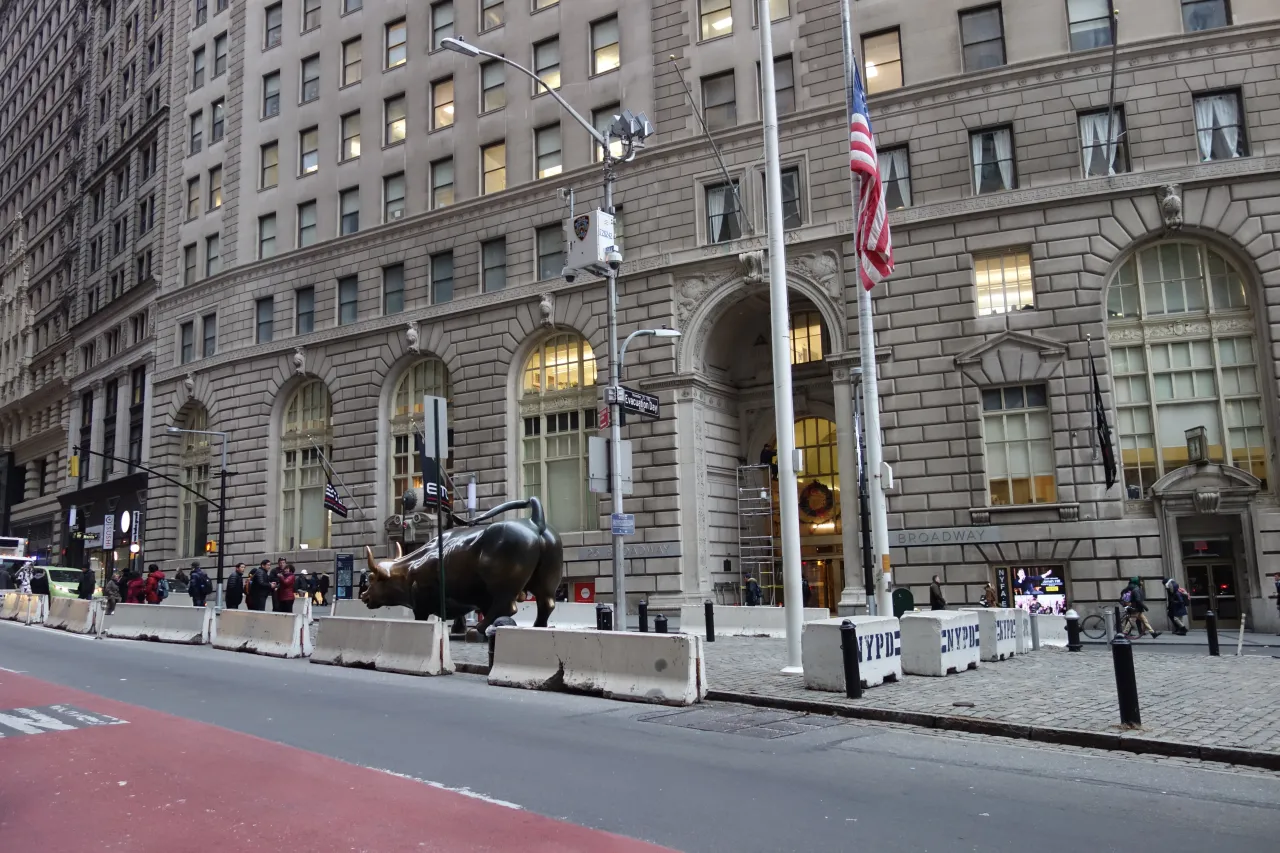 Brązowy byk na Wall Street, symbol hossy, stoi przed budynkiem z flagą USA.