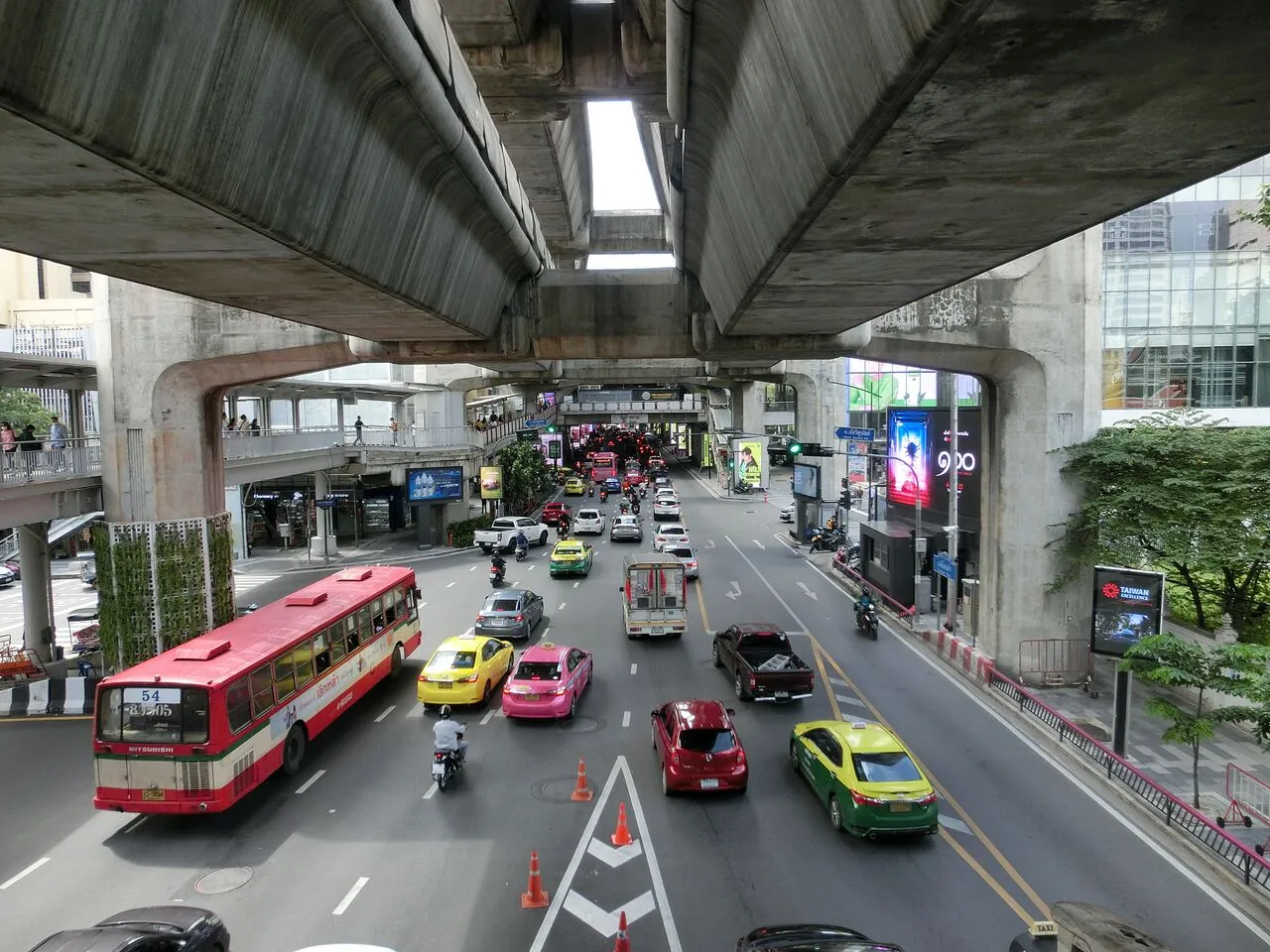 Intensywny transport w Bangkoku: czerwony autobus, żółte i różowe taksówki, samochody i motocykle pod betonową estakadą.