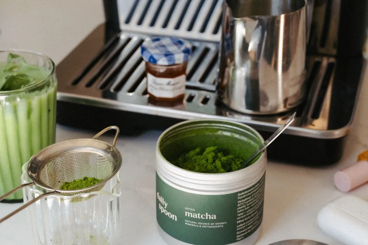 Przygotowanie herbaty matcha: proszek matcha w puszce, sitko z proszkiem, szklanka z napojem i ekspres do kawy.