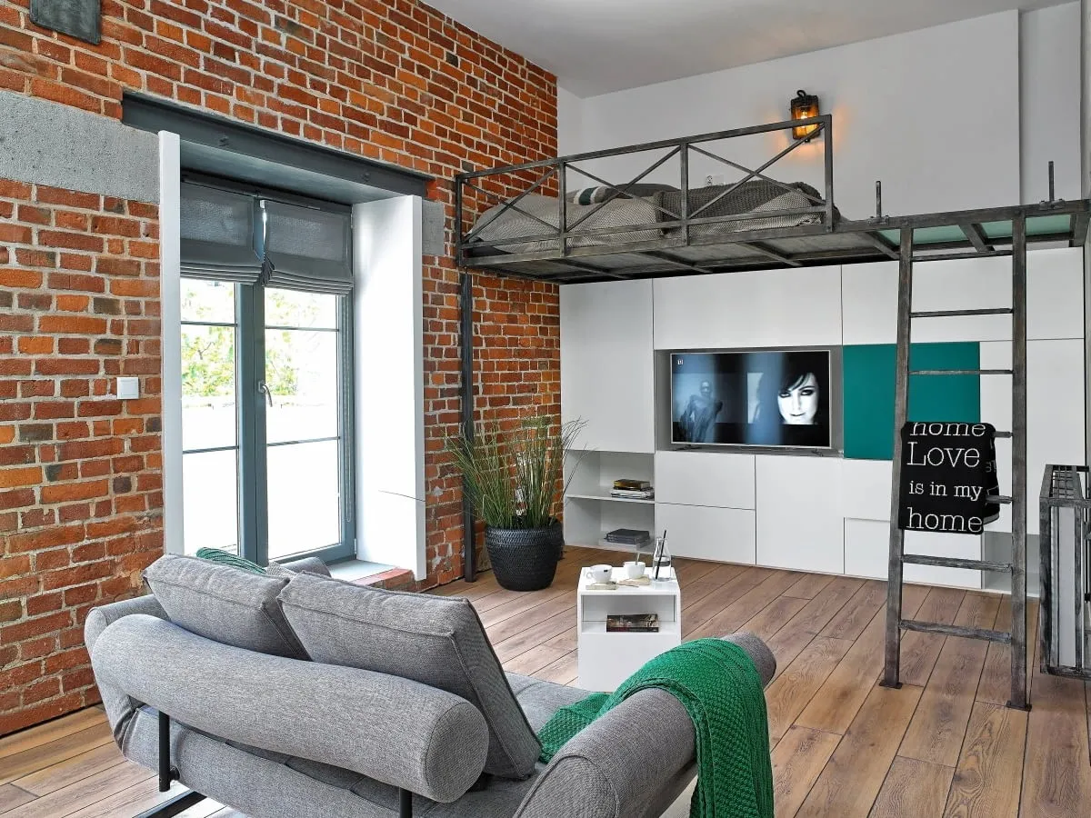 Nowoczesne mieszkanie z ceglaną ścianą, gdzie łóżko na antresoli nad szafkami RTV to sprytne rozwiązanie, co to jest antresola w mieszkaniu.