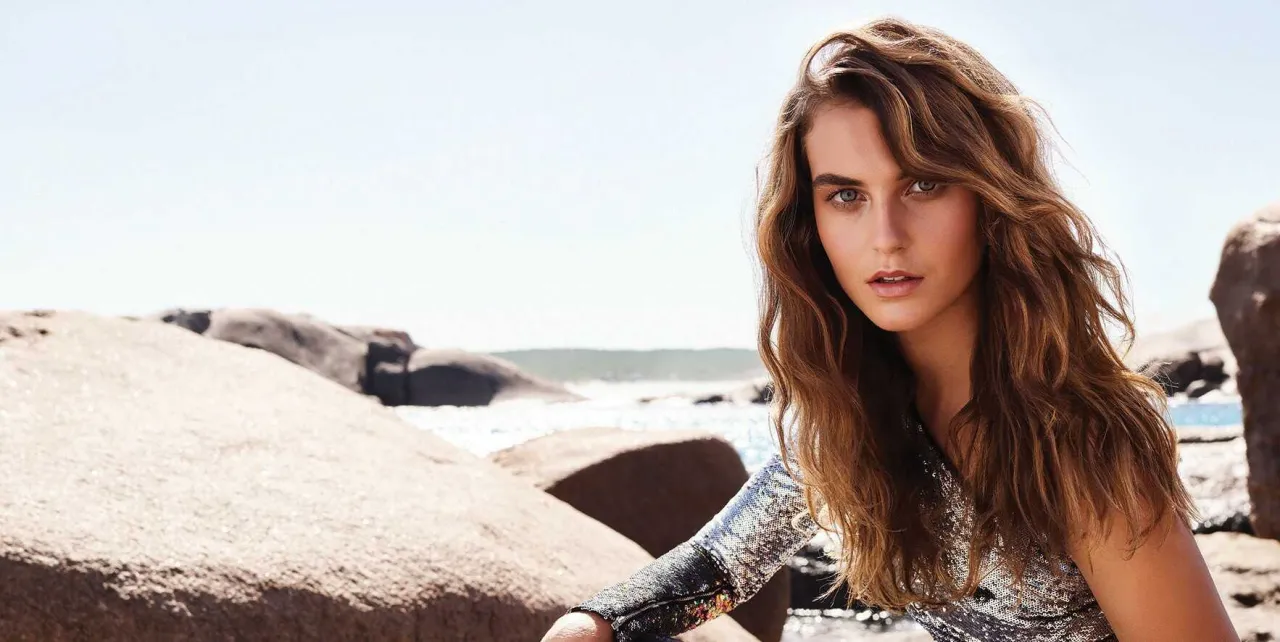 Dziewczyna z brązowymi, średnimi włosami ułożonymi w fale, siedząca na plaży.