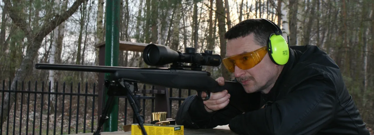 Mężczyzna w okularach i słuchawkach ochronnych celuje z karabinu. Czy myśliwi muszą brać udział w zawodach sportowych?