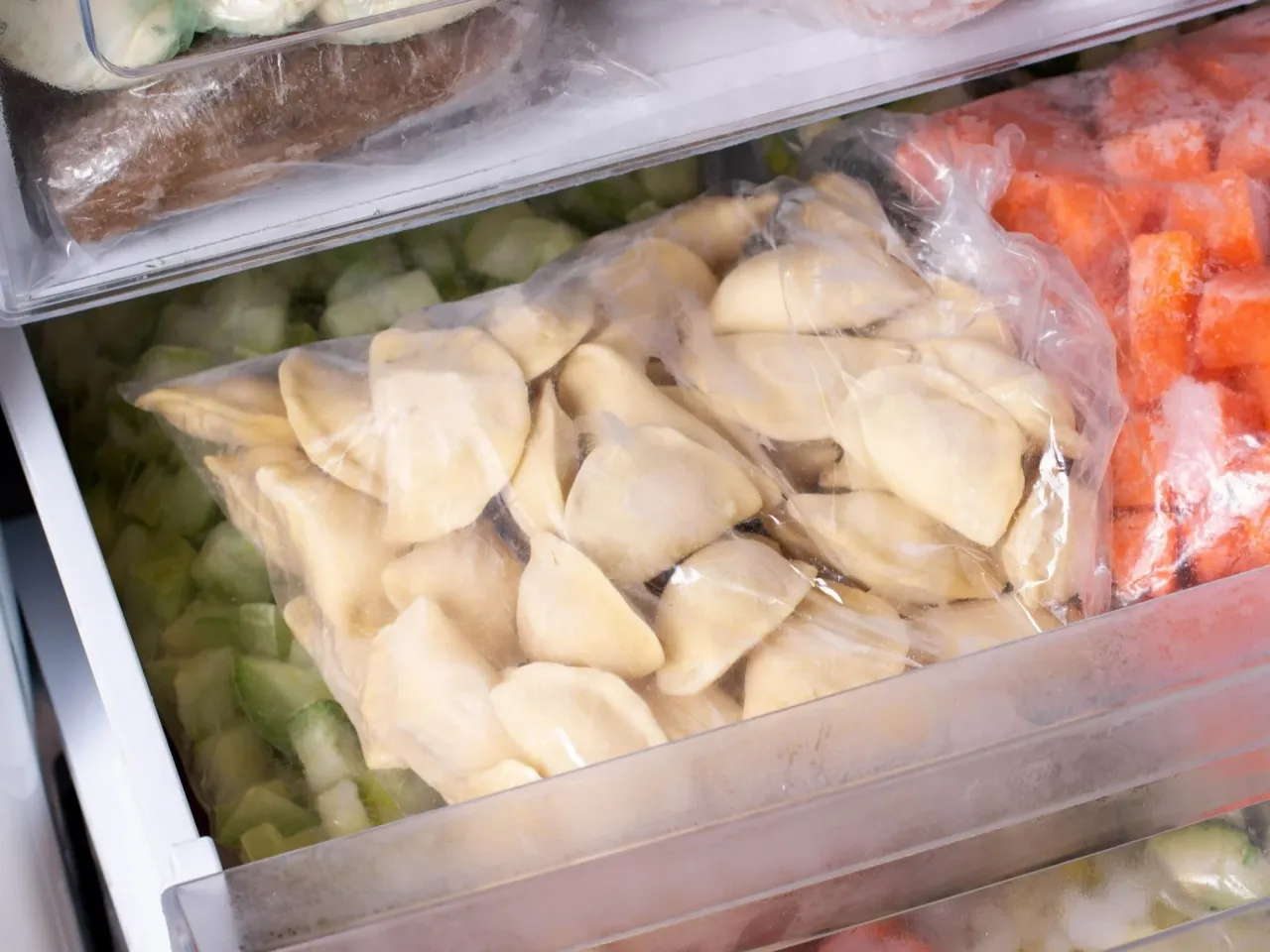 Zamrożone pierogi w woreczku, obok pokrojona marchewka i zielone warzywa. To dobry spos&oacute;b na to, jak pakować pierogi pr&oacute;żniowo, by zachowały świeżość.
